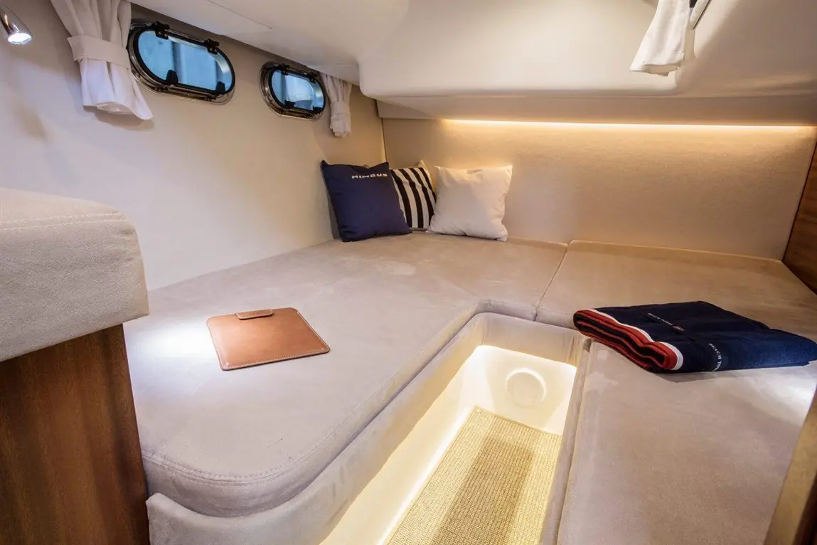 Interior of a marine cabin featuring a plush bed, two windows, cushions, a lighted storage area, and a dark towel.